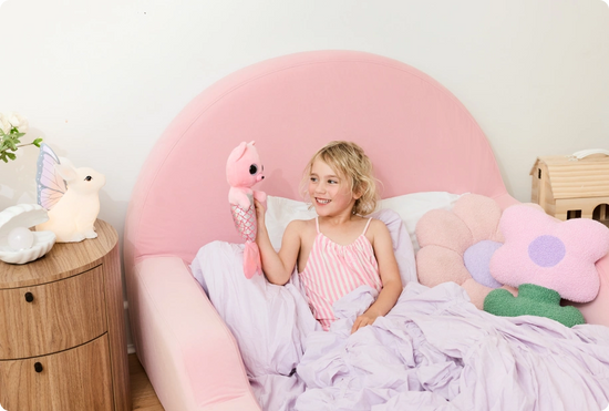 Child in a pink bed with plush toys and a nightstand.