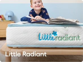 A child sitting on a bed reading a book above a white mattress or cushion with the brand name 'Little Radiant' printed in blue and green. The scene is indoors with a light blue wall and wooden furniture.